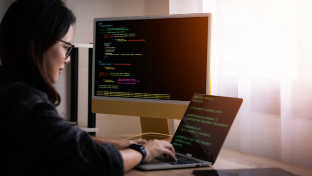 A woman is typing on a laptop computer in front of a monitor. She is wearing glasses and she is focused on her work
