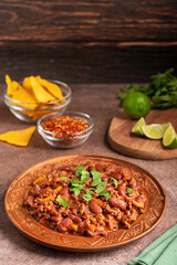 Homemade mexican spicy chili con carne stew cooked from kidney beans, tomatoes, ground beef, pepper decorated with parsley served on plate on table with sliced lime citrus and nachos tortilla chips