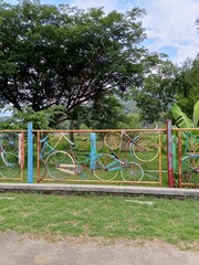 Colorful fence made from old bicycles. The unique fence is set against a backdrop of trees and lush greenery.
