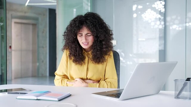 Upset young adult businesswoman suffering from stomach pain while sitting at workplace in business office. Curly brunette woman has poisoning, spasms, menstruation, constipation or digestive problems