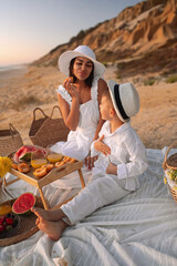 A mother and her son share a joyful picnic on a sandy beach, enjoying fresh fruits and the serene sunset. Perfect moment of family bonding and summer relaxation.