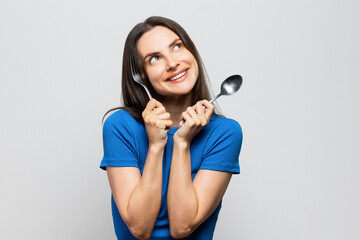 Cheerful young woman with blue top holding spoon and fork, hungry, ready to eat. On white background