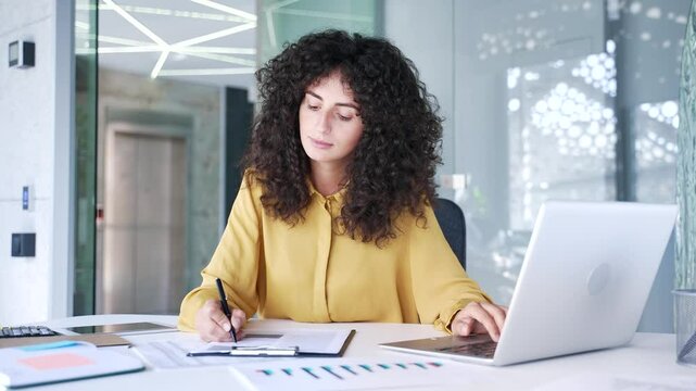 Busy young adult businesswoman is doing paperwork using laptop computer sitting at workplace in business office. Confident female financier working fills out documents, writing, engaged in accounting