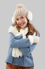 Cheerful woman embracing herself in winter Christmas time. Wearing a cozy blue sweater, pom-pom beanie hat with ear warmers, scarf, and gloves. She smiling at the camera, isolated on a gray background