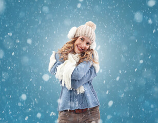Cheerful woman embracing the beauty of a winter snowfall. She is smiling at the falling snow during a white Christmas. Wearing cozy blue sweater, pom-pom beanie hat with ear warmers, scarf, and gloves