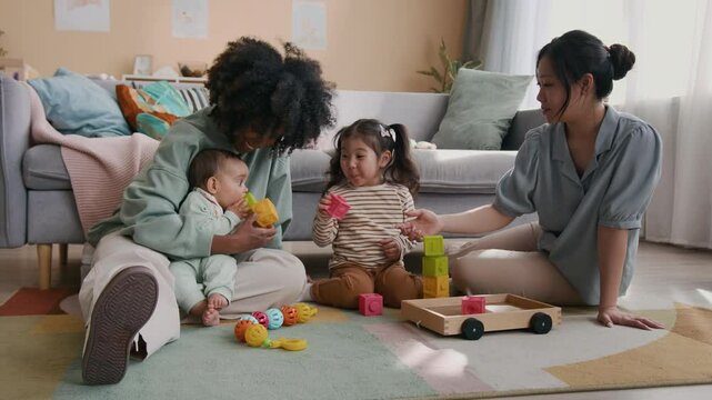 Cute little Asian girl in striped pullover and brown pants sitting on the floor between two intercultural women, playing with cubes and passing some to newborn baby boy sitting on his mother hands