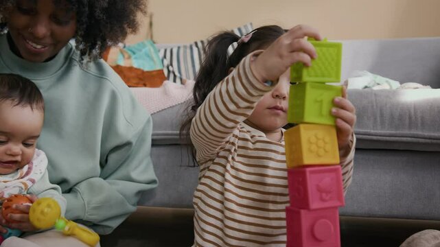 Happy Asian child in casual attire sitting by couch between her mother and young African American woman with newborn baby son on knees and building toy tower while playing with constructor