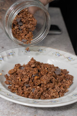 Hands pouring cereal with chocolate pieces from a glass jar into a white bowl 