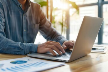 Close-up of a man typing on a laptop at a wooden desk with charts and sunlight, symbolizing productivity, work, and modern office setup.