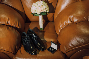 A pair of black shoes and a bouquet of flowers are on a brown couch. The shoes are placed on the couch with the bouquet of flowers in front of them. Concept of elegance and sophistication