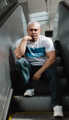 A man in a white shirt and blue jeans sits on a set of escalators. He is looking down and he is...