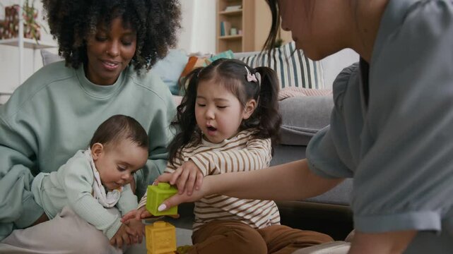 Young Asian woman giving toy cube to her little daughter sitting on the floor next to African American woman with baby son and building tower while playing with friends at home