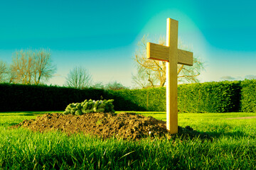 Freshly dug grave with a large, timber built golden crucifix seen in ethereal lighting. A reef is seen laid on the soil.