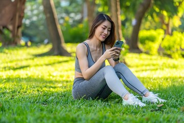 Fototapeta premium Asian Woman Sitting on Grass Checking Health Data on Smartphone and Smartwatch in Park