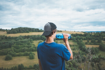 Young boy in a blue shirt and cap enjoys a refreshing drink from a water bottle while admiring the expansive green fields and rolling hills of the Beskydy Mountains