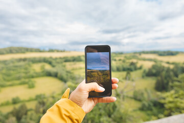 Hand holding a smartphone captures the picturesque landscape of rolling fields and green forests in the Beskydy Mountains, blending modern technology with natural beauty