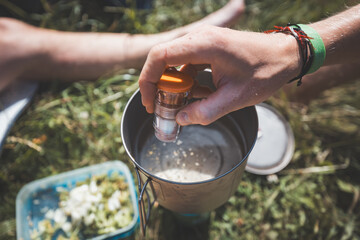 Outdoor camping scene with a person seasoning food in a portable pot, surrounded by grass, highlighting the simplicity and charm of cooking in nature