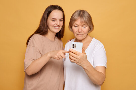 Two women share joyful moments as they explore a smartphone together in a bright yellow backdrop, showcasing technology's role in connecting generations