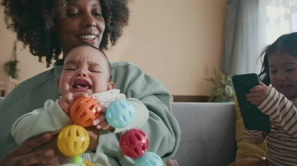Happy young African American woman sitting on couch and playing with crying baby son while little Asian girl sitting next to her and taking picture of mother and infant