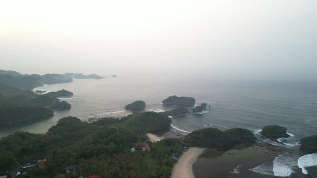 4k video footage - Aerial view of Watu Karung Beach, Pacitan, East Java, Indonesia with crashing waves during cloudy day. Cape with cliffs by the sea and green plants around at tropical island