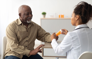 Obraz premium Pharmaceutical Drug Prescription Concept. Young black doctor showing elderly male patient pills bottle, pointing at tablets. Female specialist prescribing mature man treatment, painkiller or vitamin