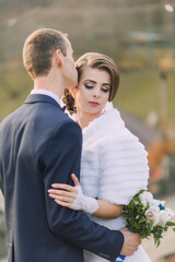 A bride and groom are embracing each other, with the bride wearing a white fur stole. The bride is holding a bouquet of flowers in her right hand. The scene is intimate and romantic
