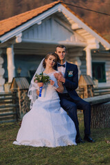 A bride and groom are sitting on a bench outside a house. The bride is holding a bouquet and the groom is holding her hand. Scene is romantic and happy