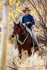 Obraz premium elderly woman in western hat and attire riding a bay Morgan mare in the fall aspen leaves in the mountains of Wyoming