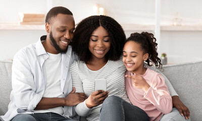 Modern Technology. Portrait of happy African American family holding and using mobile phone sitting on the couch at home. People spending time together, sharing social media, watching video or photos