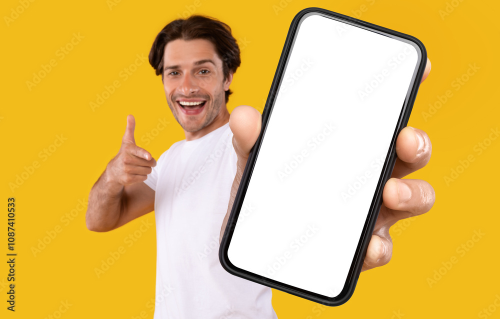 Poster cheerful young guy holding smartphone with white empty screen, pointing at blank display for mobile 