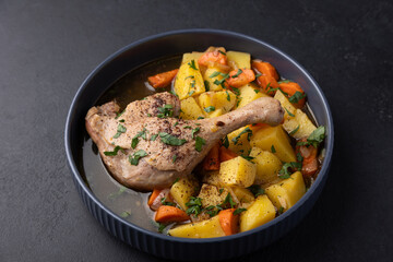 Duck leg with broth, stewed with potatoes and carrots, sprinkled with parsley and sumac. Simple home hearty cooking. Deep gray plate on a black background. Selective focus, close-up.