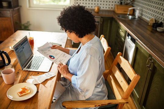 Focused woman reviewing bills and using laptop at home kitchen table