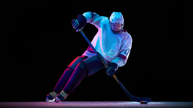 Focused man, professional hockey player in white gear in motion with stick, controlling puck against black studio background in neon light. Concept of sport, game, tournament, action, competition