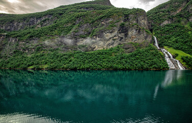 Ein Wasserfall im Geiranger Fjord rauscht in das Wasser. Spiegelung der Berge und der Wälder und...