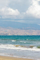 Gentle waves lapping against the sandy beach, framed by a distant mountainous coastline and a cloudy sky. Alicante, Spain. 
