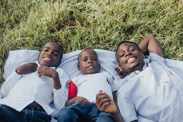 A happy family of three, wearing white shirts, lies on the grass enjoying a sunny day outdoors. They are smiling joyfully, showcasing warmth and togetherness.