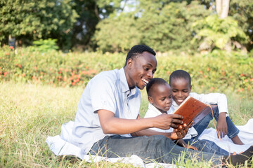 A father shares a joyful moment reading with his two sons on a sunny day in the park. The bond and happiness are highlighted by their smiles and the lush green surroundings.