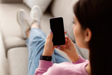 Over The Shoulder View Of Unrecognizable Young Woman Holding And Using Cell Phone, Sitting And Resting On The Couch In Living Room At Home. Selective Focus On Gadget, Template