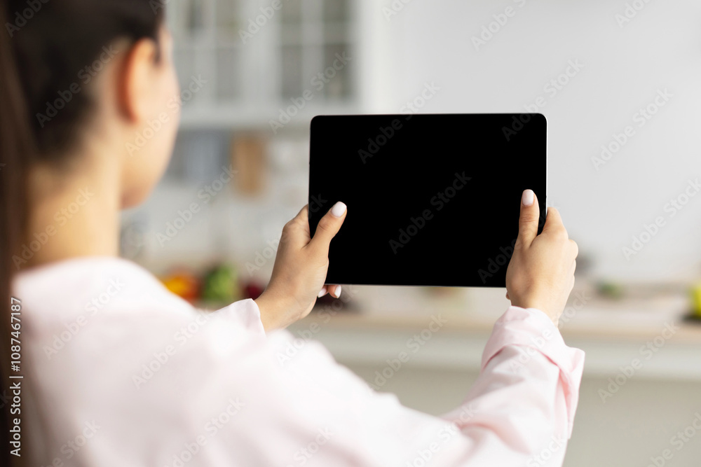 Poster Over The Shoulder View Of Unrecognizable Woman Using Digital Tablet With Empty Screen Standing In Kitchen At Home. Application Or Internet Website Advertising, Mock Up, Free Space, Selective Focus