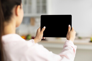 Over The Shoulder View Of Unrecognizable Woman Using Digital Tablet With Empty Screen Standing In Kitchen At Home. Application Or Internet Website Advertising, Mock Up, Free Space, Selective Focus