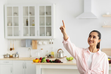 Smart Home Concept. Millennial Woman Pointing Finger Up Pushing Button On Invisible Screen Standing In Kitchen Interior Indoor. Modern Futuristic House Control System, IoT Technologies Concept