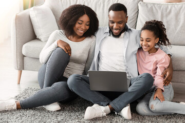 Family Weekend. Cheerful African American parents and their little daughter using laptop at home together, watching movie or browsing internet, sitting on the floor rug carpet in living room, hugging