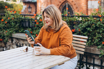 Fototapeta premium Woman holding bio coffee paper cup and smartphone, she is sitting on the cafe terrace on the city street. Urban concept. Happy Girl sitting at a table with a coffee using mobile phone.