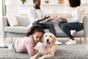 Family Conflicts. Sad little black girl hugging dog while her parents arguing in the background,...
