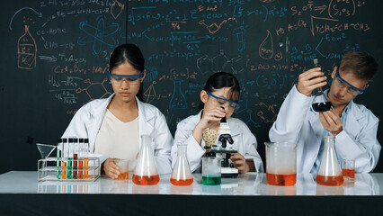 Cute girl looking under microscope while student doing experiment at blackboard with theory...