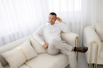 A man in a white shirt and gray pants is sitting on a couch. He is looking at the camera and he is thinking. The couch is white and has a few pillows on it