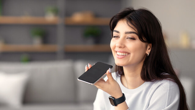 Digital Speaker Application. Portrait of young smiling woman sitting at table, talking on mobile phone, using laptop and virtual voice assistant for online search, to record reminder, dictate message