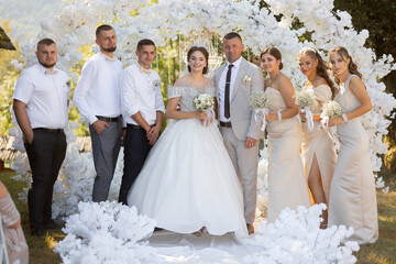 A bride and groom stand in front of a white archway with their family and friends. The bride is...