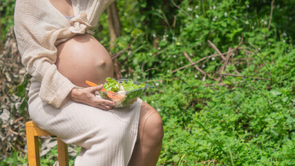 Pregnant woman holding salad bowl in hand, looking bored with eating vegetables for diet. During pregnancy On the background of natural forests and backyards.