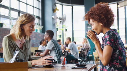 Two Young Female Friends Meeting Sitting At Table Drinking Hot Drinks Inside Coffee Shop And Talking